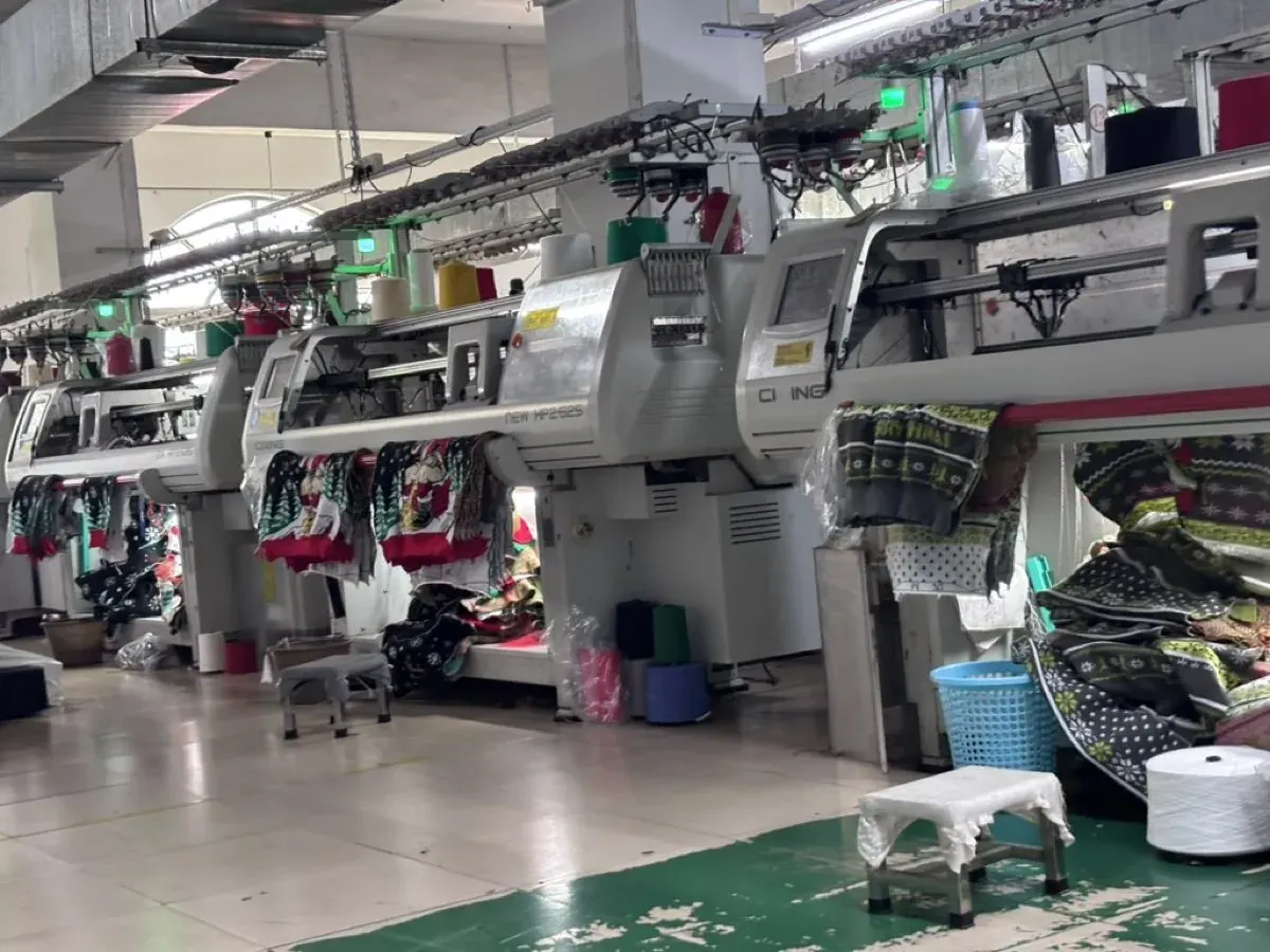 Rows of computerised flat-knit machines on our own factory floor in Dalang, with yarn cones and a clean, brightly lit workshop